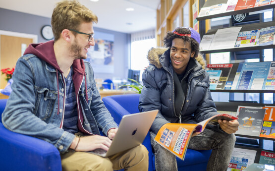 Students chatting, one holding a laptop, the other holding a magazine
