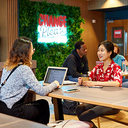 Two female students chatting in the Change Please catering outlet on Singleton Campus, one female has her laptop and the other has a cup of coffee.