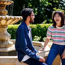 A male and female student chat and laugh sitting on the edge of the fountain at Singleton Park Abbey while enjoying the sunshine. Water is flowing from the fountain.