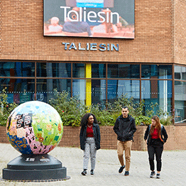 Three students walking away from the Taliesin Arts Centre on Singleton Campus. A world reimagined globe is in the foreground and you can see the Taliesin digital poster on the digital screen on the front of the building. 