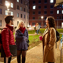 Three students chatting outside the Bay Campus Hall of Residence at Night. They are wearing coats and the area is warmly lit.