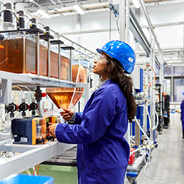 A female student is dressed in blue in a science and engineering lab. She is wearing a blue helmet and is studying some liquids in glass containers. She is also wearing goggles.
