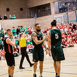 Two Swansea University basketball players shake hands during a Varsity match in the Sports Hall at Swansea Bay Sports Park. The hall is filled with supporters wearing green t shirts and red t shirts.