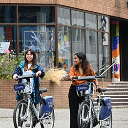 Two female students walking with Swansea University Cycles on the Mall outside the Taliesin Building on Singleton Campus. 